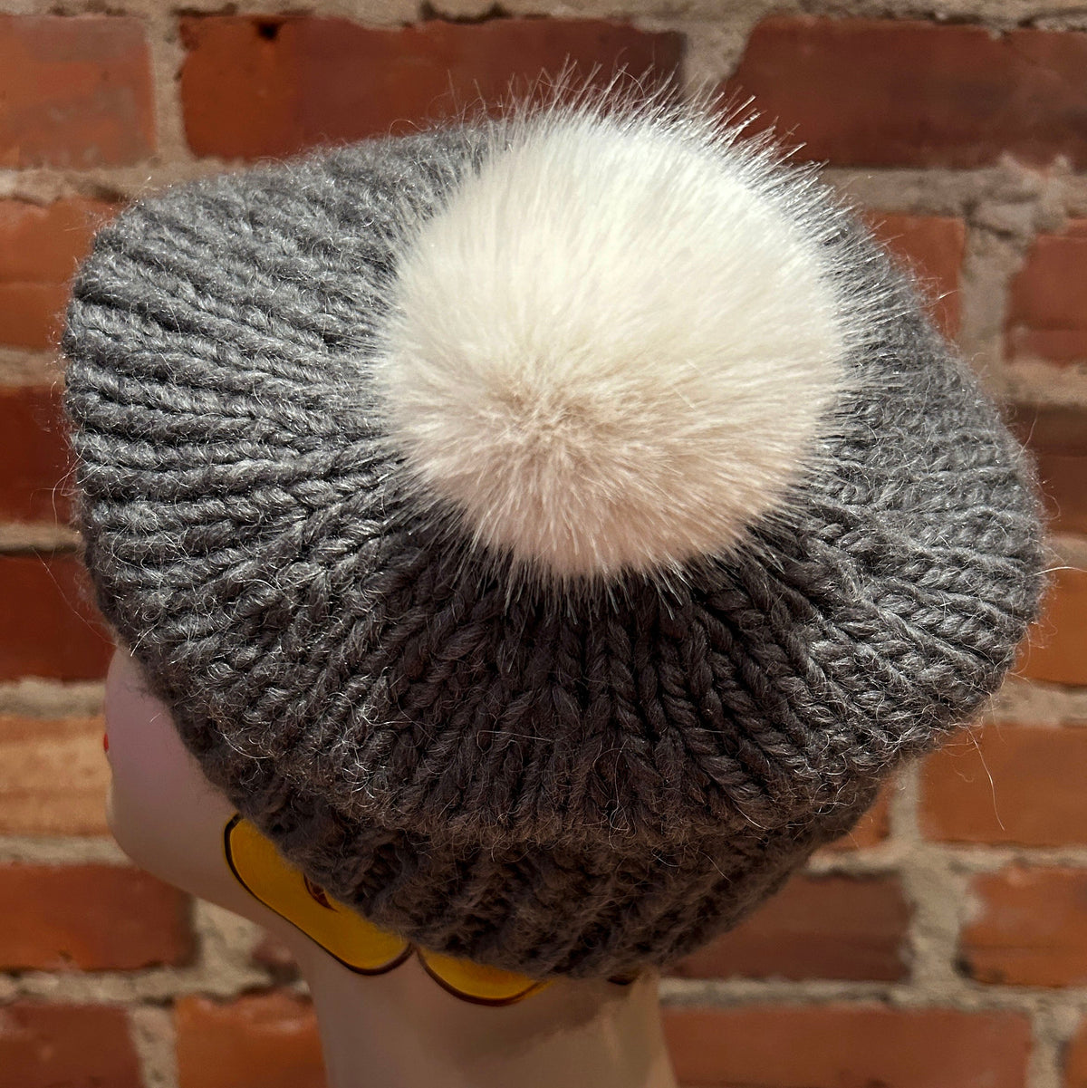 A gray ribbed knit beanie hat with a large white faux-fur pom-pom, displayed on a mannequin head against a red brick wall background.