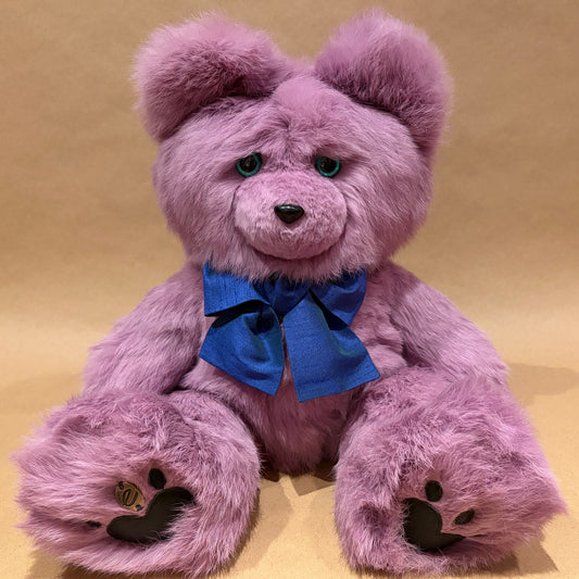A plush purple teddy bear with a blue bow around its neck, sitting against a solid tan background. The bear has green eyes, black nose and paw pads.