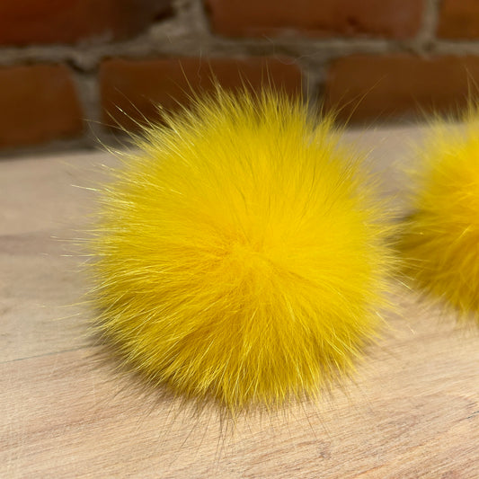 A bright yellow fluffy fox fur pom-pom resting on a light wood surface with a rustic brick wall background.