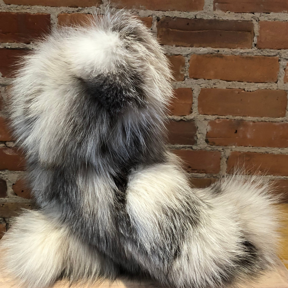 A fluffy fox fur teddy bear shown from the back against a red-brown brick wall and light wood surface.