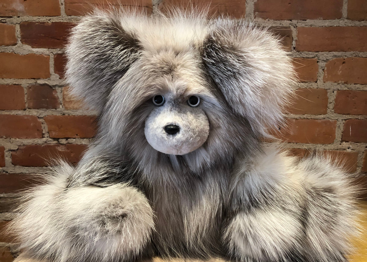 A fluffy fox-fur teddy bear with large floppy ears, a white snout, and gray-and-white fur, posed in front of a red-brown brick wall.
