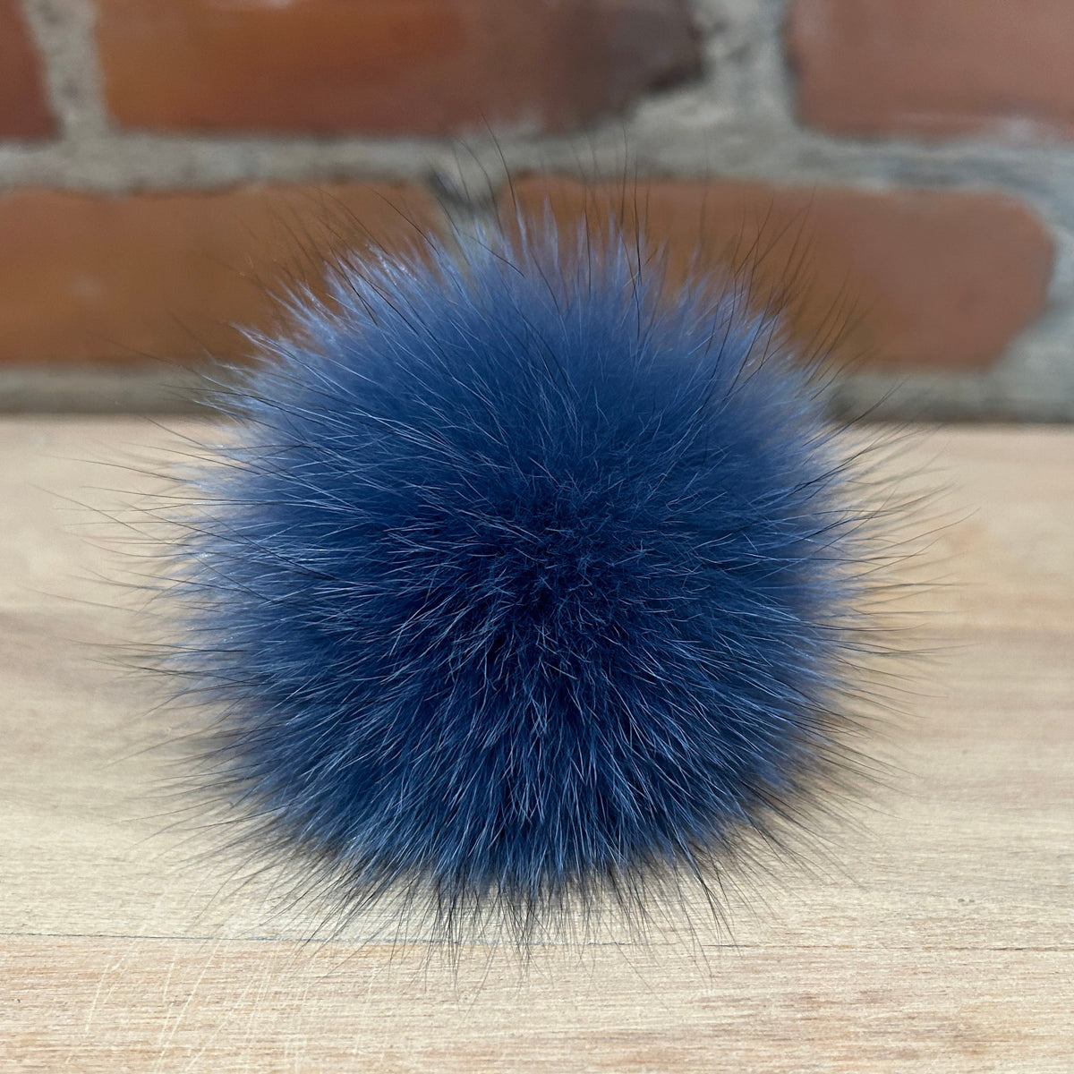 A fluffy blue fox-fur pom-pom accessory resting on a light wood surface with a blurred brick wall background.