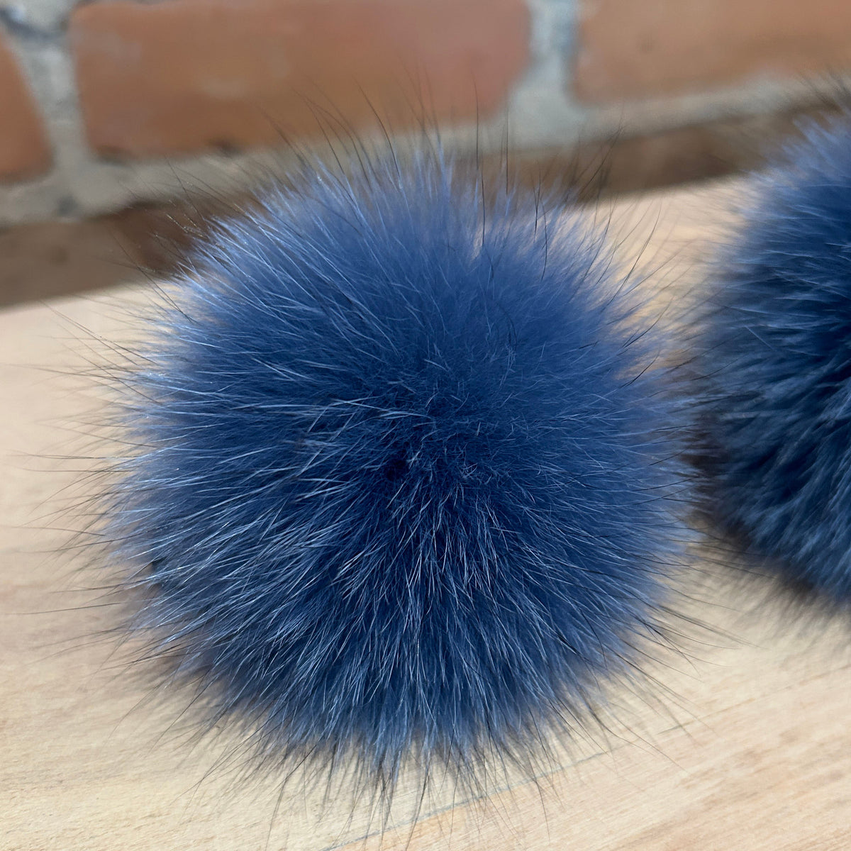 A close-up of a fluffy blue fox-fur pom pom on a light wood surface with a blurred brick wall in the background.