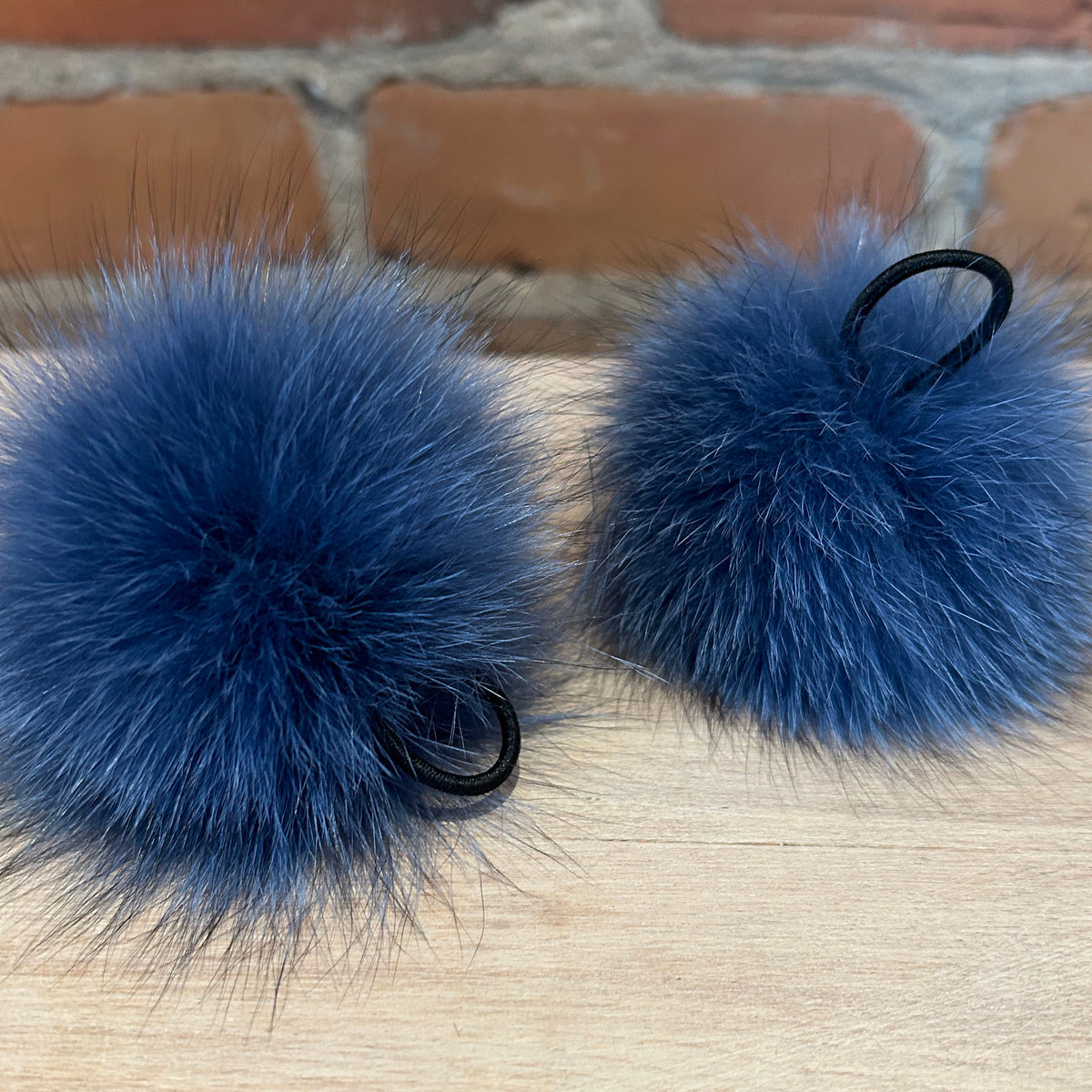 Two fluffy blue fox-fur pom-poms with black elastic loops, resting on a light wood surface with a brick wall background.