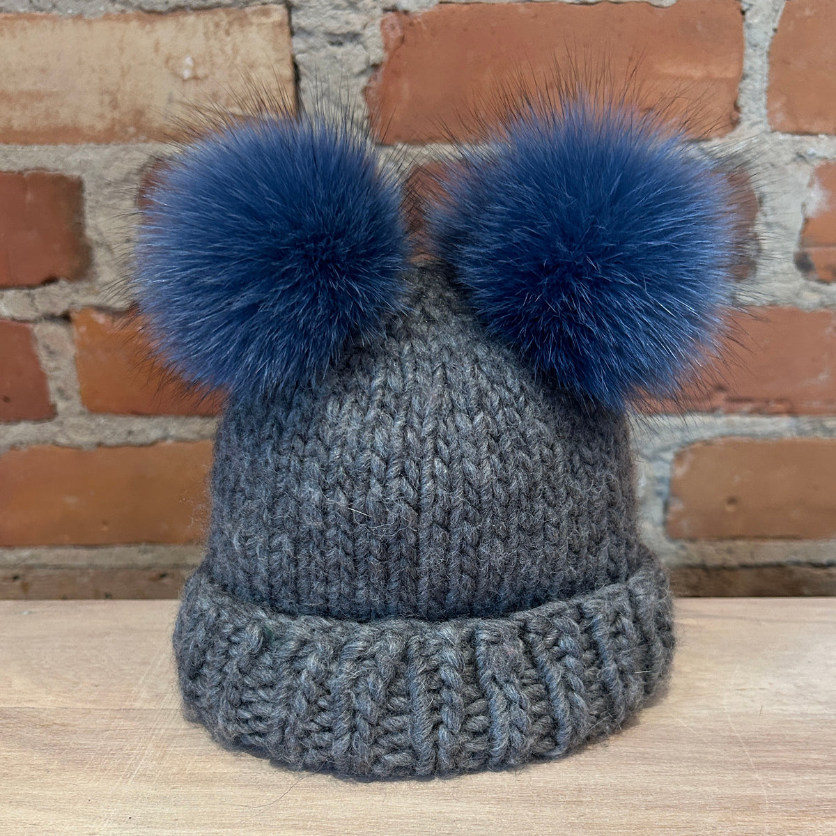 A gray knit beanie hat with two small blue fox-fur pom-poms on top, displayed on a light wood surface against a brick wall background.