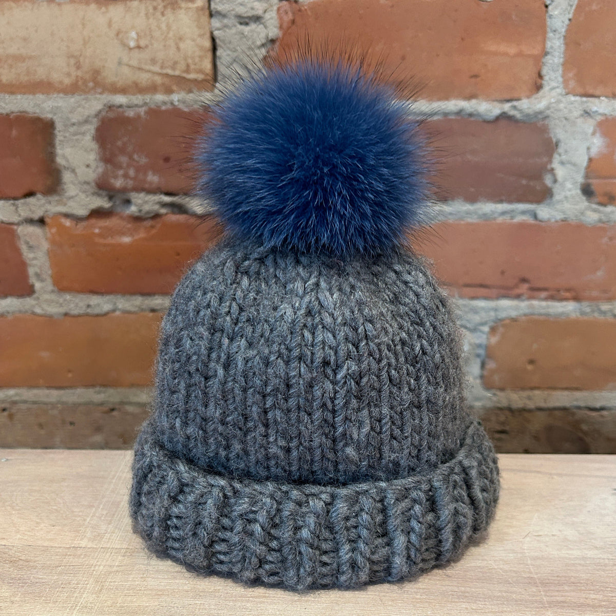 A small gray ribbed knit beanie hat with a small denim blue fox fur pom-pom, displayed on a light wood surface in front of a brick wall background.