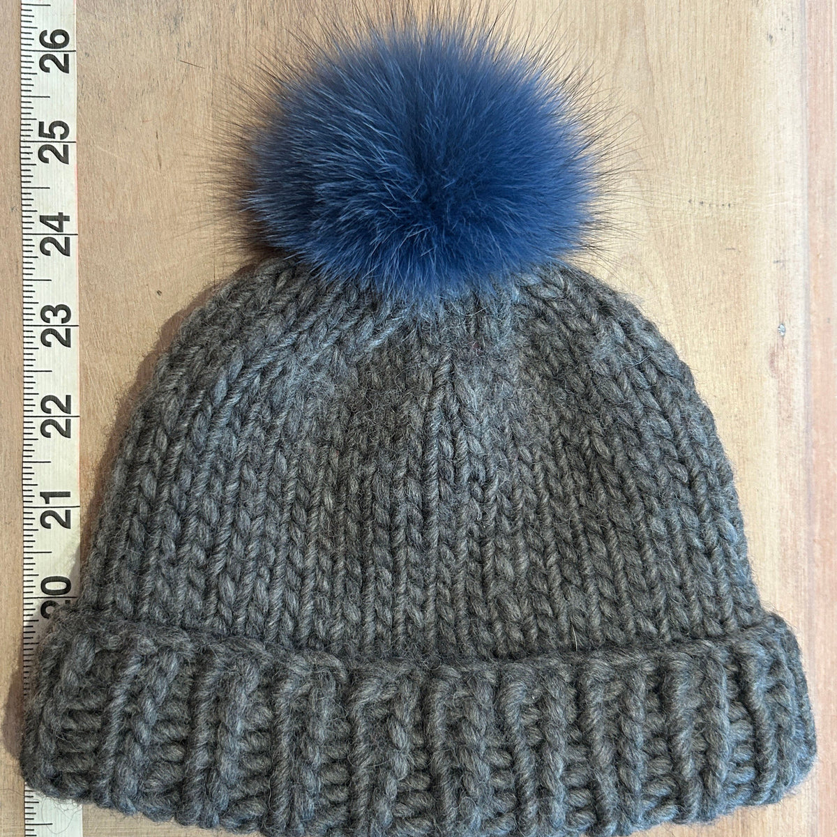 A gray ribbed knit beanie with a small blue fox fur pom-pom, shown on a light wood background with a measuring tape visible on the left side.