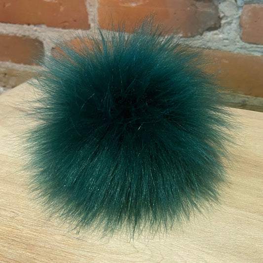 A dark clover green fluffy faux fur pom-pom accessory resting on a light wood surface with a brick wall background.
