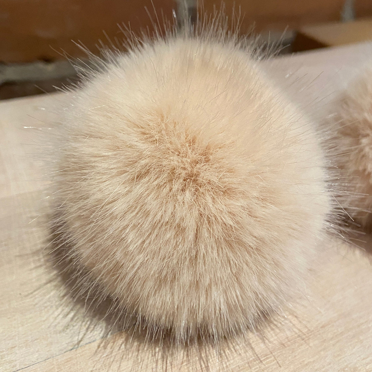 A close-up of a fluffy faux-fur pom-pom accessory in a light beige color, resting on a light wood surface with a blurred brick wall background.
