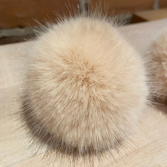 A close-up of a fluffy faux-fur pom-pom accessory in a light beige color, resting on a light wood surface with a blurred brick wall background.