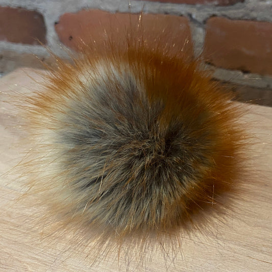 A fluffy faux-fur pom pom ball with a mix of brown, gray, and cream tones, resting on a light wood surface with a brick wall background.