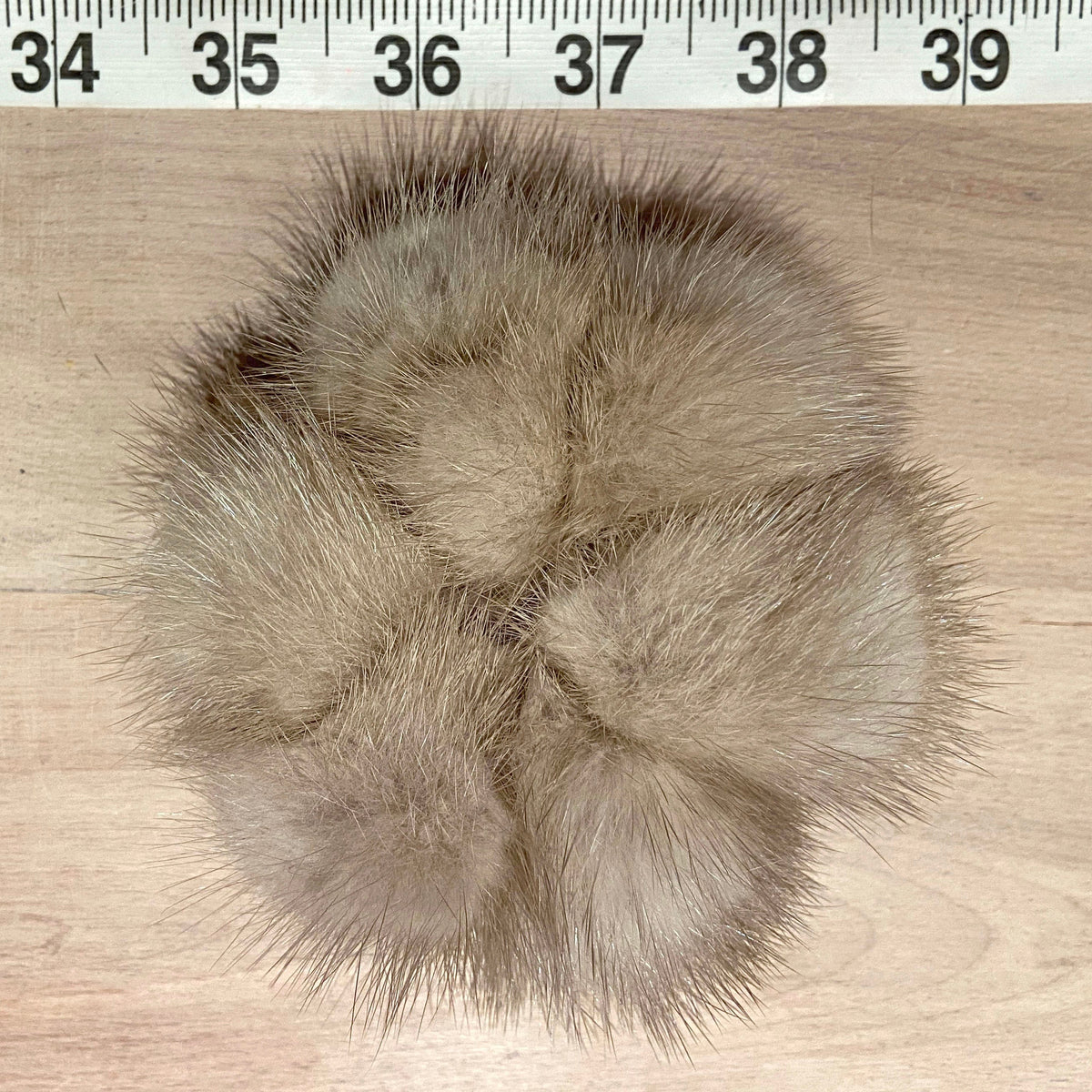 A cluster of four fluffy faux-fur pom-poms in a light taupe-brown color, shown on a light wood surface with a measuring tape ruler at the top for scale.