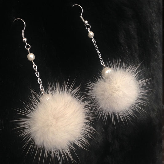 A pair of long dangle earrings with silver fishhook wires, small pearl-like beads, chain links, and fluffy cream mink-fur pom-poms, displayed against a black textured background.