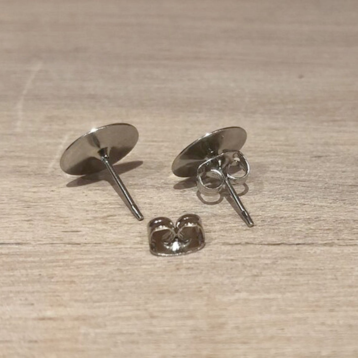 Two silver-toned flat-back post earrings with butterfly backings, displayed on a light wood surface.