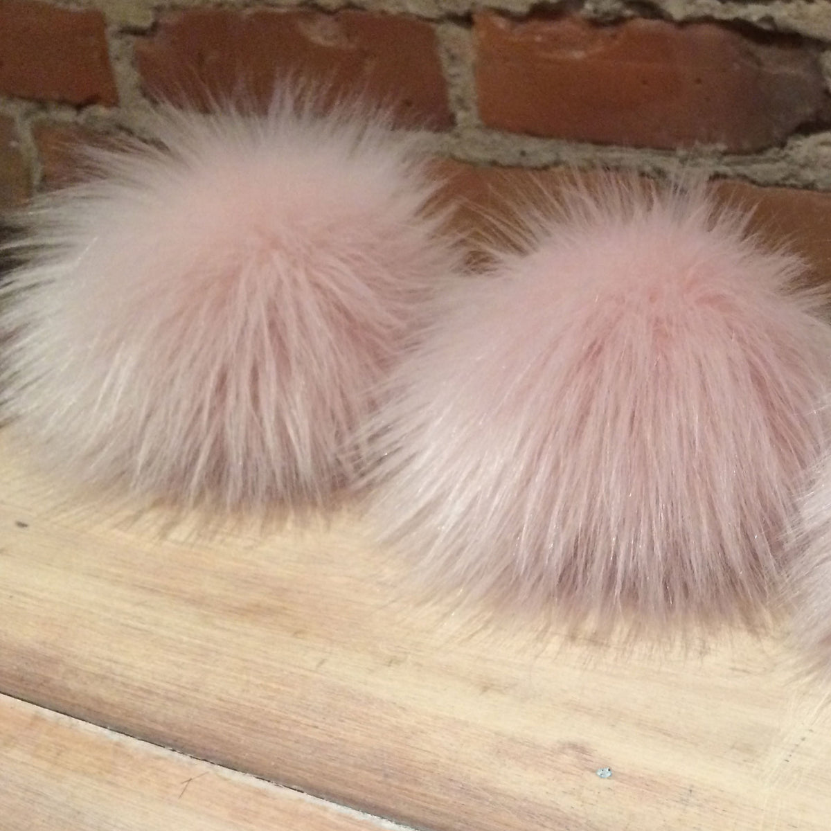 Two fluffy blush pink faux fur pom-poms resting on a light wood surface against a brick wall background.
