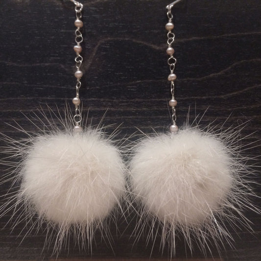 A pair of dangling earrings with silver-tone chain links, small pearl-like beads, and large fluffy white mink-fur pom-poms, displayed against a dark wood background.