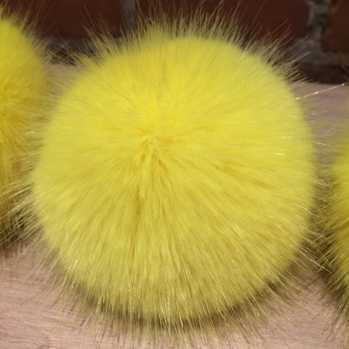 A bright yellow fluffy faux fur pom-pom ball shown in a close-up view, resting on a light wood surface.