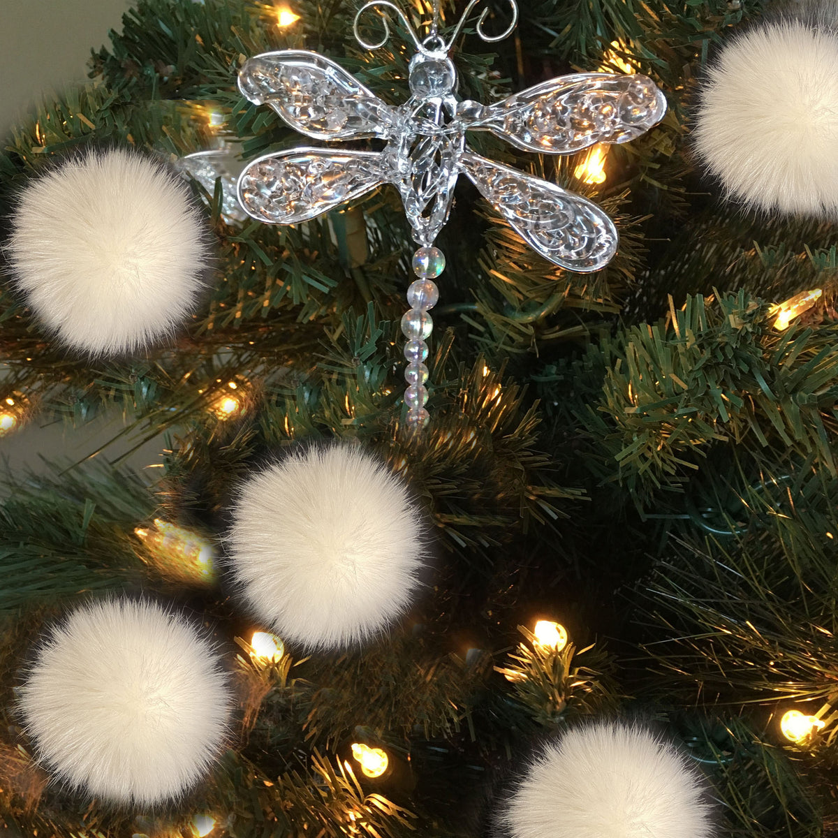 A silver dragonfly Christmas tree ornament with a beaded stem, displayed on a green artificial Christmas tree with warm white lights and white pom-poms.