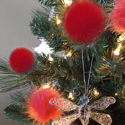 A close-up of a decorated Christmas tree with three bright red fluffy pom-pom ornaments, warm white string lights, and a clear glittery dragonfly ornament.