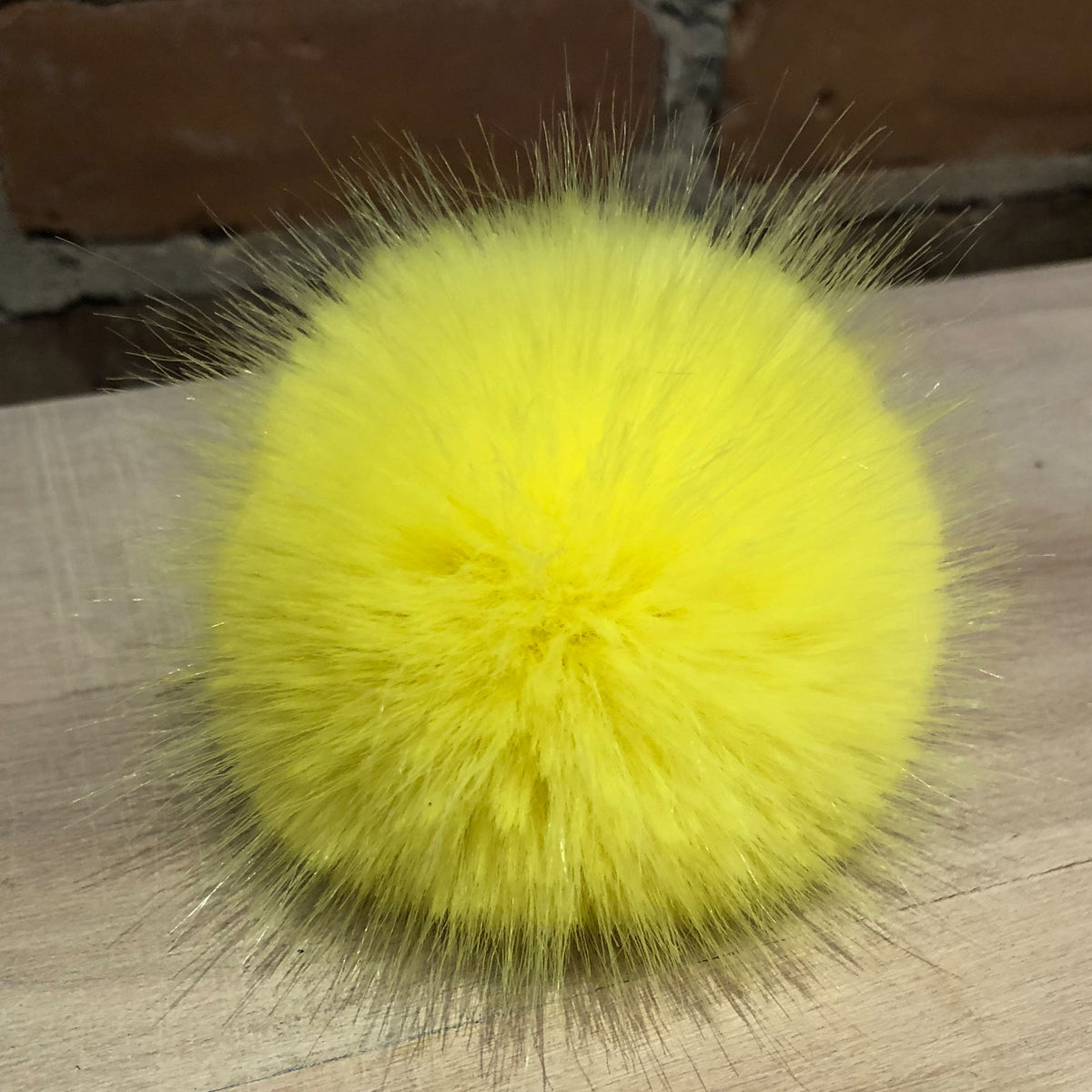 A bright yellow faux fur pom-pom with long, wispy fibers, resting on a light-colored wooden surface with a brick wall in the background.