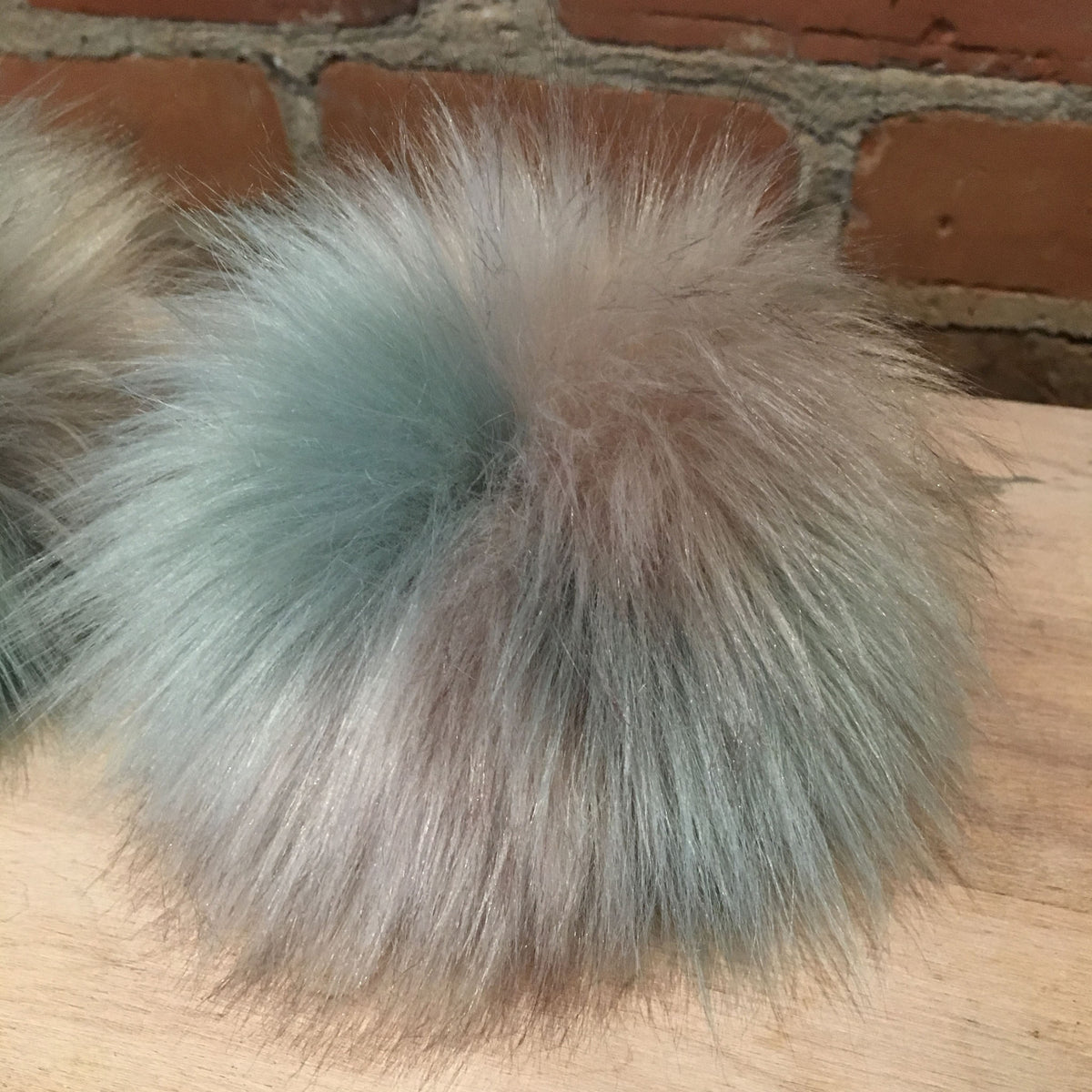 A fluffy faux-fur pom pom ball in a soft blend of pale blue, beige, and gray tones, resting on a light wood surface with a brick wall in the background.