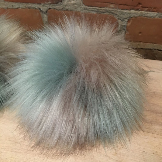 A fluffy faux-fur pom pom ball in a soft blend of pale blue, beige, and gray tones, resting on a light wood surface with a brick wall in the background.