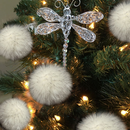 A clear, ornate dragonfly Christmas ornament with iridescent bead details, displayed on a green artificial Christmas tree with warm white lights and fluffy white pom-poms.