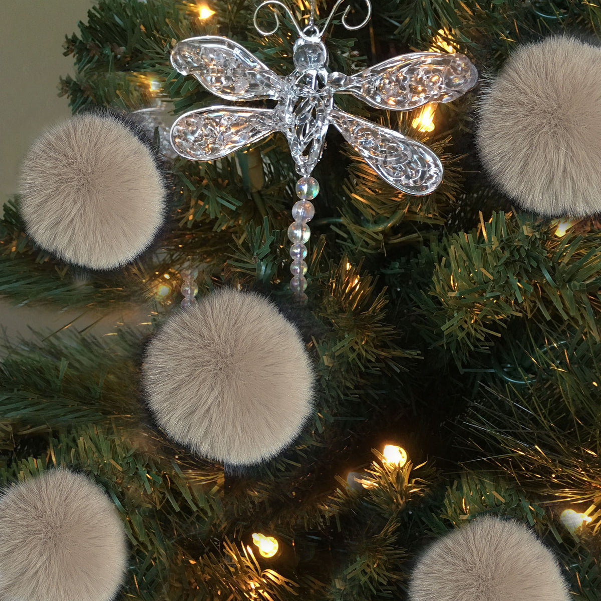 A silver dragonfly Christmas tree ornament with a beaded tail, displayed on a green artificial Christmas tree with warm white lights and beige faux fur pom-poms.