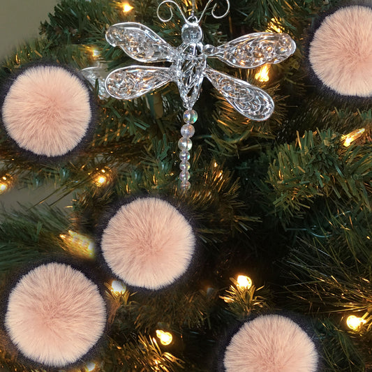 A silver dragonfly Christmas tree ornament hanging on a green artificial tree with warm white lights, surrounded by pink faux-fur pom-pom ornaments.