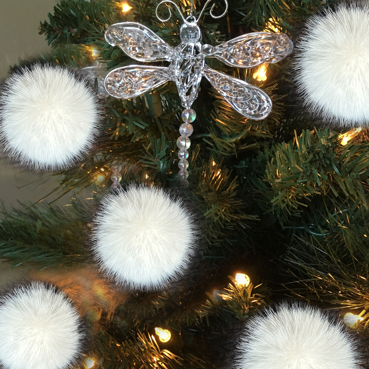 A clear, ornate dragonfly Christmas ornament hanging on a green artificial Christmas tree with warm white lights and fluffy white pom-pom ornaments around it.