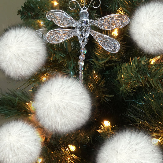 A clear, ornate dragonfly Christmas ornament with a beaded tail, displayed on a green artificial Christmas tree with warm white lights and fluffy white pom-pom ornaments.