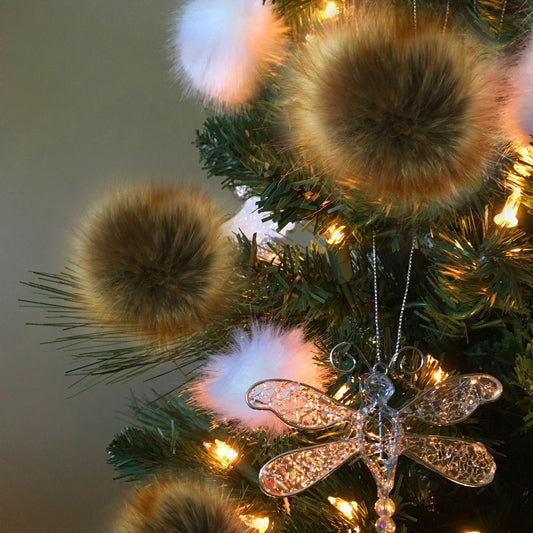A close-up of a decorated Christmas tree with large brown faux-fur pom-poms, soft white fluffy ornaments, warm string lights, and a clear glitter dragonfly ornament.
