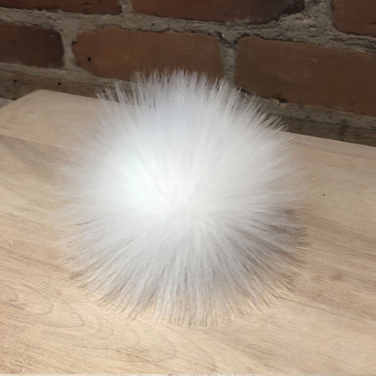 A fluffy white faux fur pom-pom accessory resting on a light wood tabletop with a brick wall background.