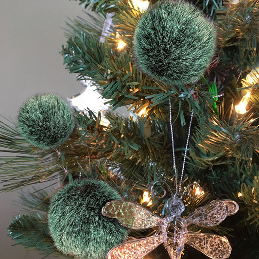 Three dark green fuzzy pom-pom Christmas ornaments hanging on artificial green tree branches with warm white string lights, alongside a glittery dragonfly ornament.