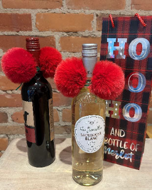 Two wine bottles decorated with bright red fluffy pom-poms, standing in front of a brick wall with a red plaid gift bag that reads 'HO HO HO and a bottle of Merlot'.