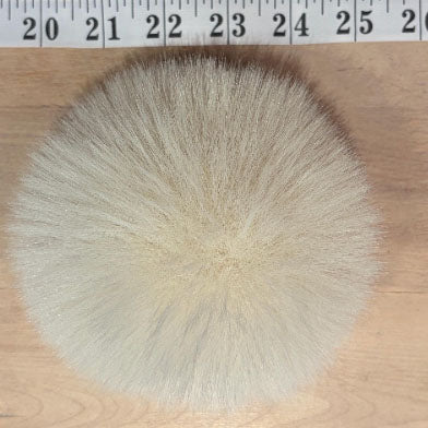 A round, fluffy faux fur pom-pom in a light cream or off-white color, shown on a light wood surface with a measuring tape ruler at the top to indicate scale.