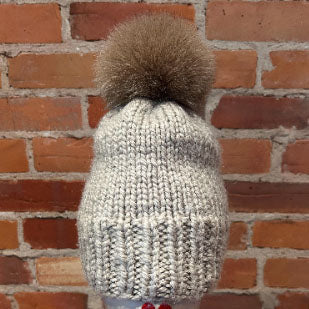 A chunky knit beanie hat in a light oatmeal gray color with a large gold fox fur pom-pom, displayed against a red brick wall background.