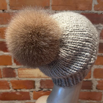 A chunky knit beanie in light gray with a large fluffy gold fox-fur pom-pom, displayed on a mannequin head against a red brick wall background.