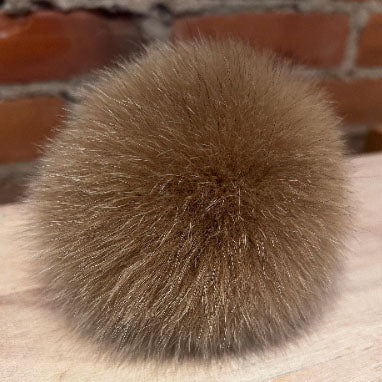 A round, fluffy pom-pom ball in a warm brown color, resting on a light wooden surface with a blurred brick wall in the background.