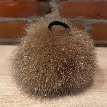 A fluffy brown fox fur pom-pom hair tie accessory with a black elastic band, resting on a light wood surface with a blurred brick wall background.