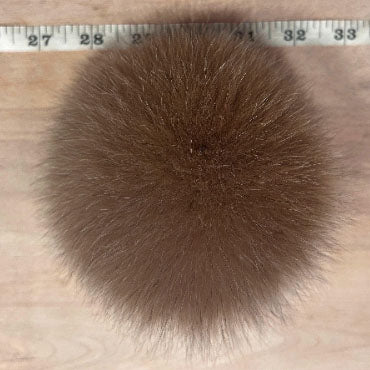 A round golden brown fur pom-pom accessory shown from above on a light wood surface, with a measuring tape visible along the top edge for scale.
