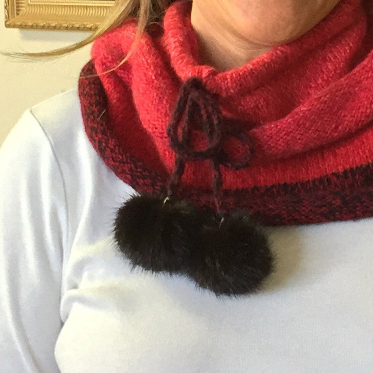 A close-up of a person wearing a red knit cowl scarf with a dark patterned edge and two black mink-fur pom-poms hanging from the front.