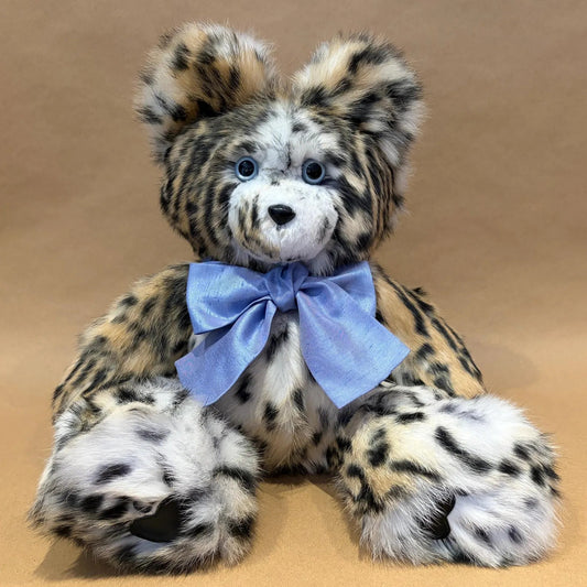 A plush leopard or snow leopard teddy bear with black and tan spots on a white and tan base, wearing a light blue satin bow around its neck, set against a solid tan background.