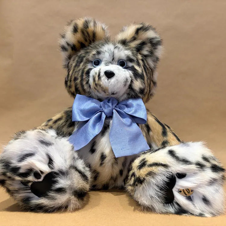 A plush leopard-style teddy bear with black and tan spots, a white muzzle, blue eyes, and a light blue satin bow around the neck, posed against a plain tan background.