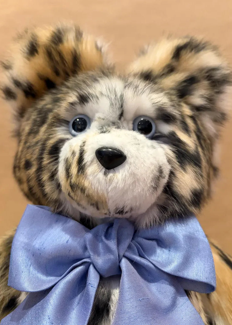 A close-up of a plush teddy bear with mottled tan, black, and white fur, large dark eyes, and a light blue satin bow tie against a warm beige background.