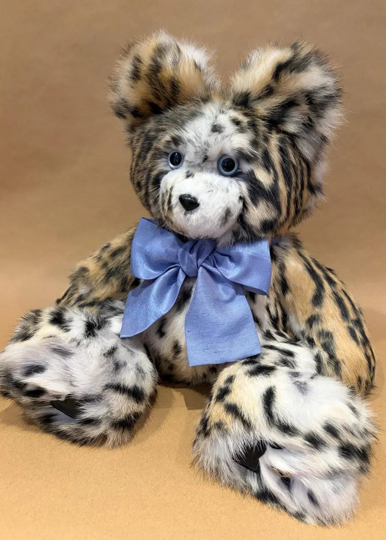 A plush teddy bear with spotted leopard-like fur in tan, black, and white, wearing a light blue satin bow, posed against a plain tan background.