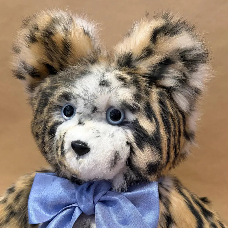 A close-up of a plush teddy bear with spotted tan, black, and cream fur, blue eyes, and a large light blue satin bow around its neck, set against a plain tan background.
