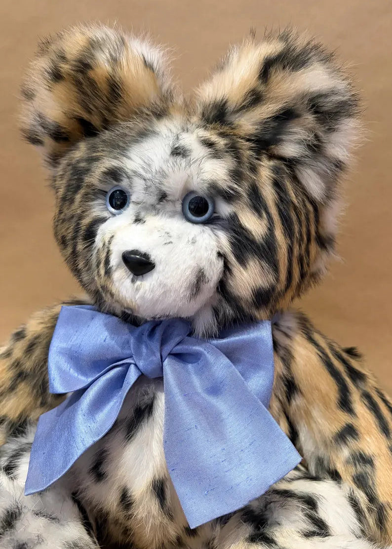 A close-up of a plush teddy bear with spotted tan, black, and cream fur, blue eyes, and a large light blue satin bow around its neck, set against a plain tan background.