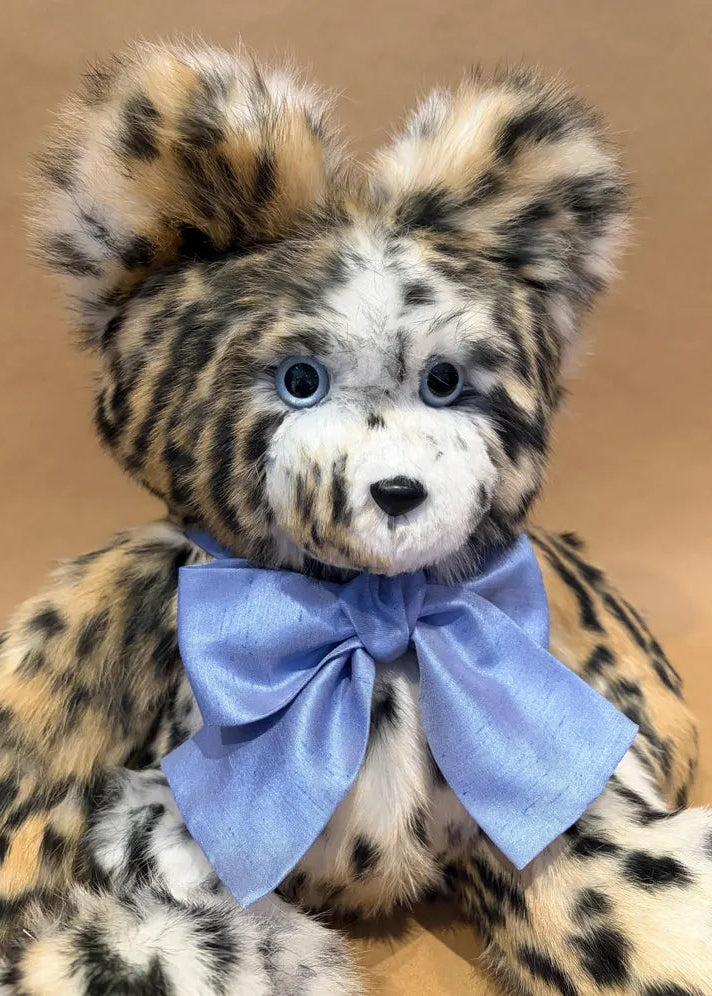 A plush leopard-style teddy bear with tan, black, and cream fur, blue eyes, and a light blue satin bow around its neck, photographed against a plain warm beige background.