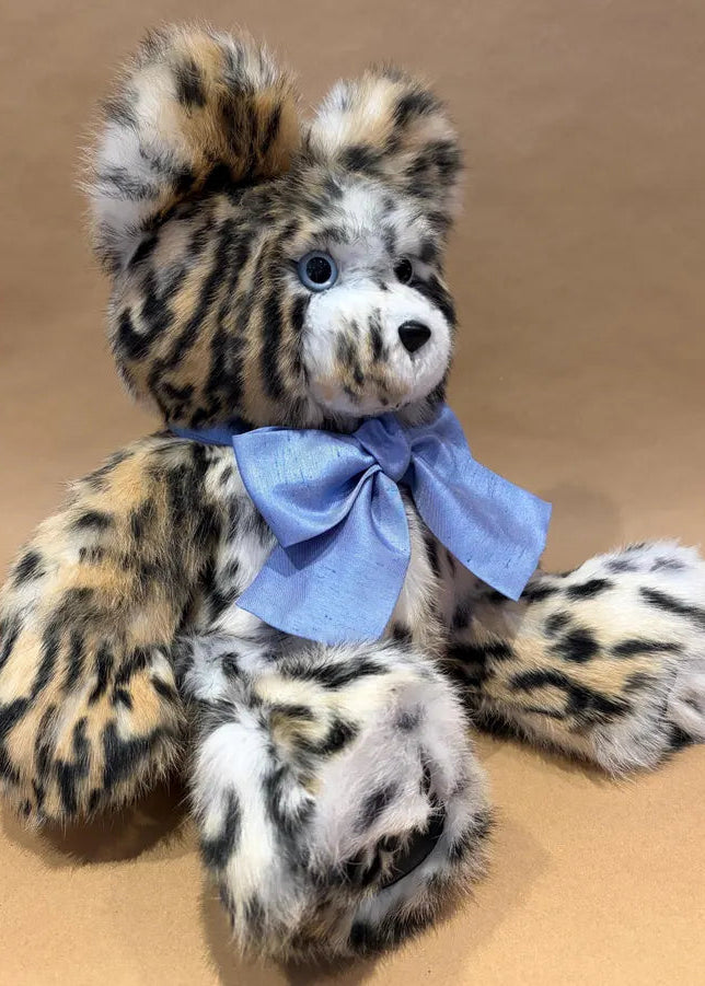A plush teddy bear with a spotted animal-print pattern in black, beige, and white, wearing a light blue satin bow around its neck, set against a plain tan background.